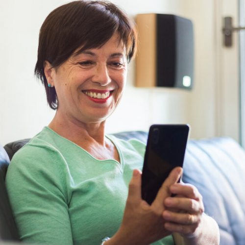 Mature,Older,Woman,Smiling,Using,Smartphone,Sitting,On,The,Sofa